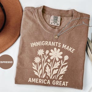 以下が含まれることがあります： 「IMMIGRANTS MAKE AMERICA GREAT」の文字と花の模様が入ったエスプレッソ色のTシャツ。ペンダント付きのシルバーのネックレスがシャツの上に置かれています。シャツは折りたたまれ、白い表面に表示されています。