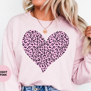 May include: Light pink sweatshirt featuring a large heart design with a pink and black leopard print pattern. The sweatshirt is a soft, casual style, perfect for everyday wear. The text "LIGHT PINK" is in a pink circle in the bottom left corner.