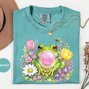 May include: Seafoam green t-shirt with a colorful graphic of a frog blowing a bubblegum bubble. The frog is surrounded by flowers and a bee. A gold necklace with a star pendant is on the shirt.