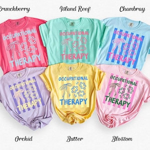 Könnte beinhalten: Sechs farbige T-Shirts in Rosa, Blau, Grün, Gelb und Lila. Jedes Shirt zeigt den Text "OCCUPATIONAL THERAPY" in einem vertikal gestreiften Design mit einer Schleife und Puzzleteilen.