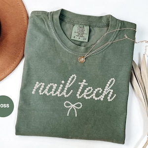 May include: A moss green t-shirt with the words "nail tech" spelled out in white pearl-like beads. A small bow made of similar beads is below the text. A gold necklace and a brown hat are also in the image.