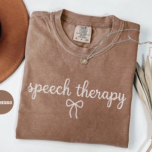 May include: Espresso-colored t-shirt with the words "speech therapy" in white script and a bow graphic. A silver necklace with a pendant rests on the shirt. The shirt has a Comfort Colors tag.
