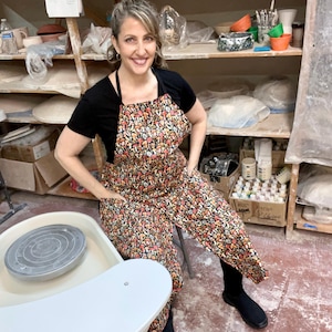 Może przedstawiać: Kobieta w czarnym t-shircie i długim czarnym fartuchu z nadrukiem grzybów. Fartuch ma dwie kieszenie. Stoi w pracowni ceramicznej z kołem garncarskim na pierwszym planie.