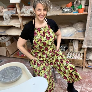 May include: A woman wearing a black shirt and a green, red, and yellow floral apron stands in a pottery studio. She is standing in front of a pottery wheel.
