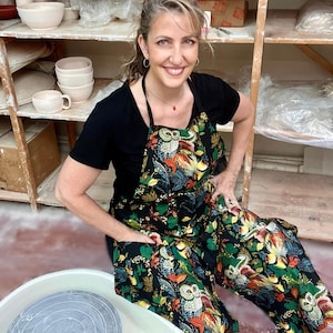 May include: A black apron with a colorful owl and floral pattern. The apron is worn by a person in a pottery studio. The apron has a pocket on the front. Pottery wheels and shelves with ceramic bowls are in the background.