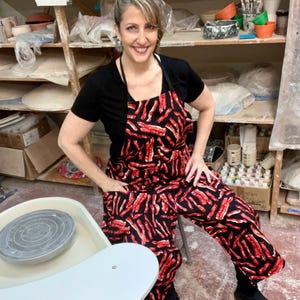 May include: A woman wearing a black apron with a bacon print sits in front of a pottery wheel. The apron has large pockets on the front. The woman is smiling and looking at the camera.