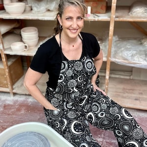 Puede incluir: Una mujer lleva un delantal negro con un estampado celestial blanco, con lunas, estrellas y soles. Está de pie en un estudio de cerámica, con estantes de cerámica en el fondo.