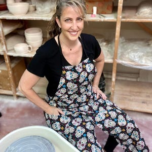May include: A woman wearing a black t-shirt and a black apron with a colorful sugar skull pattern. She is sitting in front of a pottery wheel.