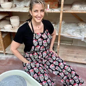 May include: A woman wearing a black apron with a floral and sugar skull pattern. The apron has a large pocket on the front. She is standing in front of a pottery wheel.