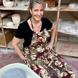 May include: A woman wearing a black t-shirt and a colorful apron with a skull and rose pattern. She is sitting in front of a pottery wheel.