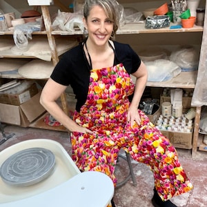 May include: A woman wearing a black top and a colorful floral apron and pants. The apron and pants feature a vibrant pattern of pink, yellow, and orange flowers. A pottery wheel is in the foreground.