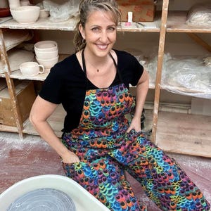 May include: A woman wearing a black t-shirt and a colorful apron and pants with a peacock feather design. The apron and pants have a vibrant pattern of blue, green, orange, and yellow. Pottery and shelves are in the background.