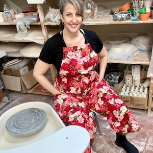 May include: A woman wearing a black t-shirt and a red floral apron stands in front of a pottery wheel. The apron has a large floral pattern with pink and red roses.