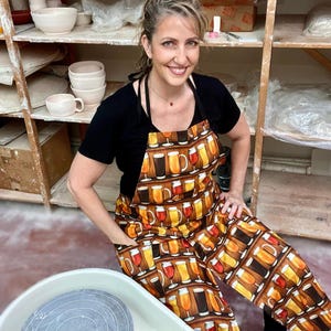 May include: A woman wearing a black t-shirt and a brown apron with a pattern of beer glasses. She is sitting in front of a pottery wheel.