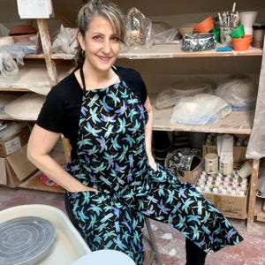 May include: A woman wearing a black apron with a dragonfly print. The apron has two pockets. The woman is standing in a pottery studio.