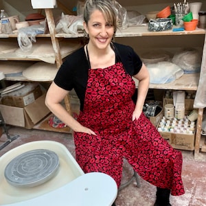 May include: A red apron with a black lip print is worn over a black top. The apron has pockets and is worn by a person in a pottery studio. Shelves in the background hold pottery supplies and finished pieces. A pottery wheel is in the foreground.