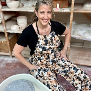 May include: A woman wearing a black shirt and a dog-print apron is sitting in a pottery studio. She is smiling and looking at the camera. The apron has a variety of dog breeds on it, including golden retrievers, labs, and poodles.