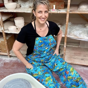 May include: A blue apron with a sea turtle pattern. The apron has a black neck strap and pockets. The person in the image is smiling and sitting in a pottery studio. Shelves with pottery are in the background.