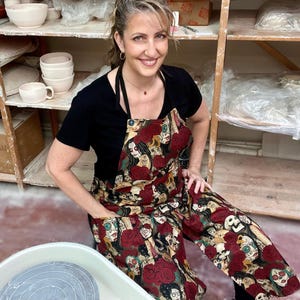 May include: A woman wearing a black t-shirt and a red apron with a Day of the Dead print. The apron has a large pocket on the front. She is sitting in front of a pottery wheel.