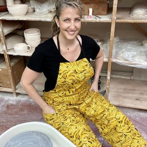 May include: A woman wearing a black t-shirt and a yellow apron and pants with a banana pattern. The woman is smiling and sitting near a pottery wheel. Shelves with pottery items are in the background.