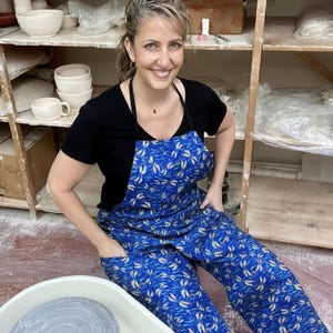 May include: A woman wearing a blue apron with a white and gold leaf pattern, black shirt, and black apron straps. The apron has pockets. Ceramic bowls and shelves are in the background.