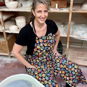 May include: A woman wearing a black shirt and a colorful skull-patterned apron is sitting in front of a pottery wheel. She is smiling and looking at the camera. There are shelves of pottery behind her.