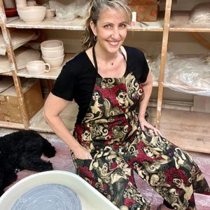 May include: A woman wearing a black t-shirt and a black apron with a colorful Day of the Dead print. The apron features images of skulls, flowers, and women. She is sitting in a pottery studio with a pottery wheel in front of her.