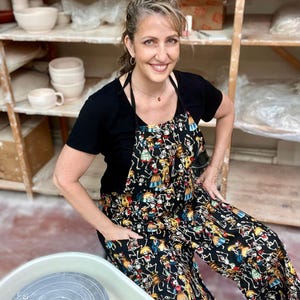 May include: A woman wearing a black apron with a colorful skeleton pattern. The apron has a large pocket on the front. She is standing in a pottery studio with shelves of pottery in the background.