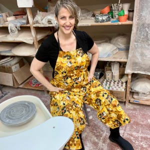 May include: A woman wearing a yellow and brown apron with a cat print stands in a pottery studio. She is wearing a black t-shirt and black shoes. A pottery wheel is in the foreground.