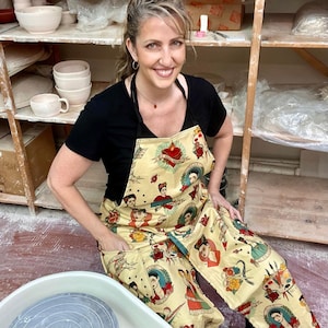 May include: A woman wearing a black t-shirt and a yellow apron with a colorful Frida Kahlo print. The apron has pockets and a heart with a dagger design. She is sitting in front of a pottery wheel.