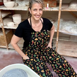 May include: A woman wearing a black apron and pants with a colorful Mexican-themed pattern featuring sombreros, cacti, and maracas. The woman is smiling in a pottery studio, with shelves of ceramic bowls and mugs in the background.