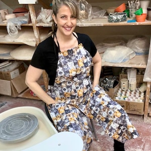 May include: A woman wearing a black top and a cat-print apron. The apron features a repeating pattern of various cat breeds in shades of gray, brown, and white. The woman is smiling in a pottery studio, with shelves of pottery and supplies in the background.
