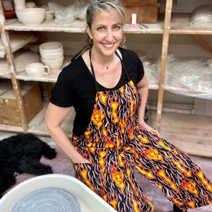 May include: A woman wearing a black t-shirt and a black and yellow apron with a flame pattern. She is standing in a pottery studio with a pottery wheel in the foreground.