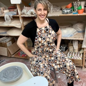 May include: A woman wearing a black shirt and a brown and white apron with a coffee cup pattern. She is standing in front of a pottery wheel and shelves filled with pottery.