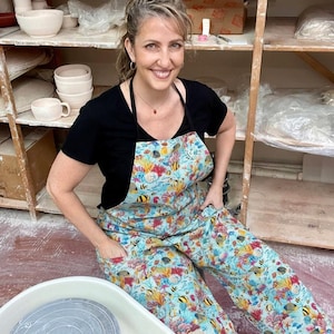 May include: A woman wearing a black t-shirt, a colorful apron, and matching pants. The apron and pants feature a vibrant underwater scene with coral, fish, and other marine life. She is sitting in a pottery studio with shelves of ceramic bowls.