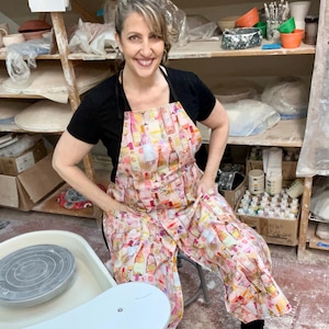 May include: A woman wearing a pink and white apron with a pattern of wine bottles. She is standing in a pottery studio with a pottery wheel in the foreground.