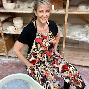 May include: A woman wearing a black apron with a colorful Day of the Dead print. The apron features skulls, flowers, and women with sugar skull makeup. She is standing in a pottery studio.