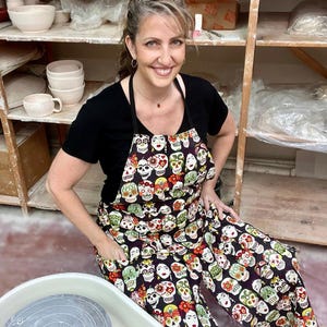 May include: A woman wearing a black t-shirt and a black apron with a colorful sugar skull pattern. She is standing in a pottery studio with shelves of pottery in the background.