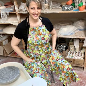 May include: A woman wearing a colorful floral apron with a pottery wheel in the foreground. The apron has a variety of colors and patterns, including green, blue, yellow, and pink. The pottery wheel is white and has a gray spinning plate.