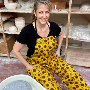 May include: A woman wearing a black shirt and a yellow apron and pants with a sunflower pattern. She is smiling and sitting in a pottery studio. Shelves in the background hold white pottery.