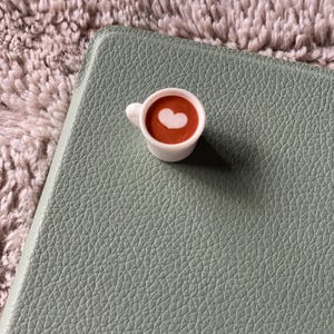 Pode incluir: Uma pequena x&iacute;cara de caf&eacute; branca com um desenho em forma de cora&ccedil;&atilde;o na espuma, cheia de um l&iacute;quido castanho, colocada sobre uma superf&iacute;cie texturizada verde claro. O fundo &eacute; um material macio e texturizado castanho claro.