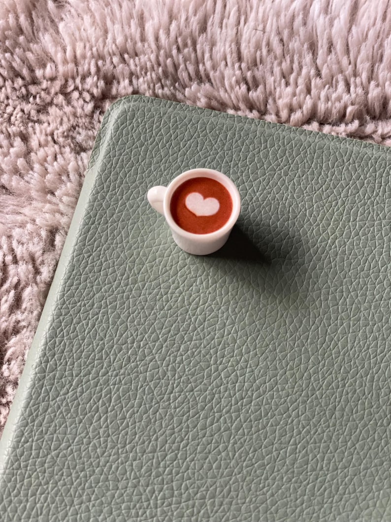 Pode incluir: Uma pequena x&iacute;cara de caf&eacute; branca com um desenho em forma de cora&ccedil;&atilde;o na espuma, cheia de um l&iacute;quido castanho, colocada sobre uma superf&iacute;cie texturizada verde claro. O fundo &eacute; um material macio e texturizado castanho claro.