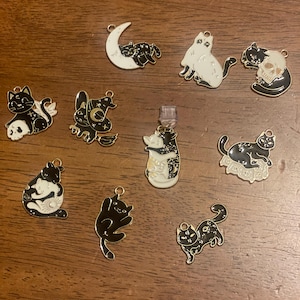Puede incluir: Una colección de diez colgantes con temática de gatos en varias poses y diseños. Los colgantes presentan esmalte negro y blanco con contornos dorados. Algunos colgantes incluyen lunas, calaveras y motivos celestiales. Medidas en centímetros.