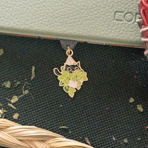 Puede incluir: Un marcapáginas dorado con un gato negro asomándose a una planta verde frondosa. El gato tiene un pecho blanco y una nariz blanca.