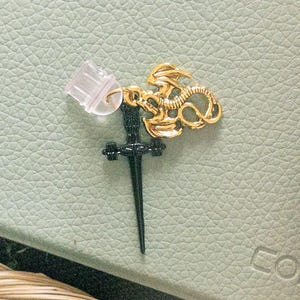 Pode incluir: Um amuleto de dragão dourado com uma adaga preta presa a ele. O amuleto está sobre uma superfície de couro verde claro.