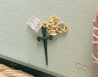 May include: A gold dragon charm with a black dagger attached to it. The charm is on a light green leather surface.