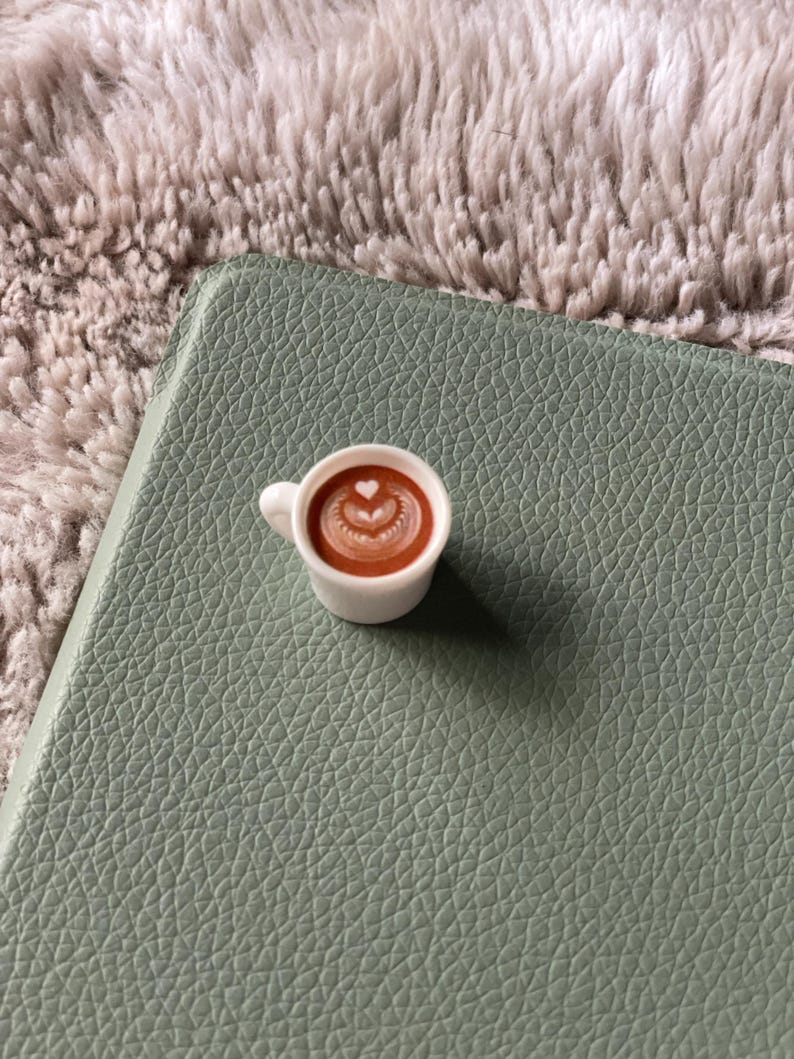 Pode incluir: Uma pequena ch&aacute;vena de caf&eacute; de cer&acirc;mica branca com um desenho em forma de cora&ccedil;&atilde;o na espuma, pousada numa superf&iacute;cie texturizada verde-s&aacute;lvia. A ch&aacute;vena est&aacute; cheia de um l&iacute;quido castanho, e o fundo &eacute; um material macio e fofo, cinzento claro.