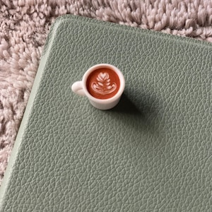 Pode incluir: Uma pequena x&iacute;cara de caf&eacute; branca com um design de arte latte, colocada sobre uma superf&iacute;cie texturizada verde s&aacute;lvia. O caf&eacute; &eacute; marrom e a arte latte &eacute; branca. O fundo &eacute; um material macio e difuso, marrom claro.