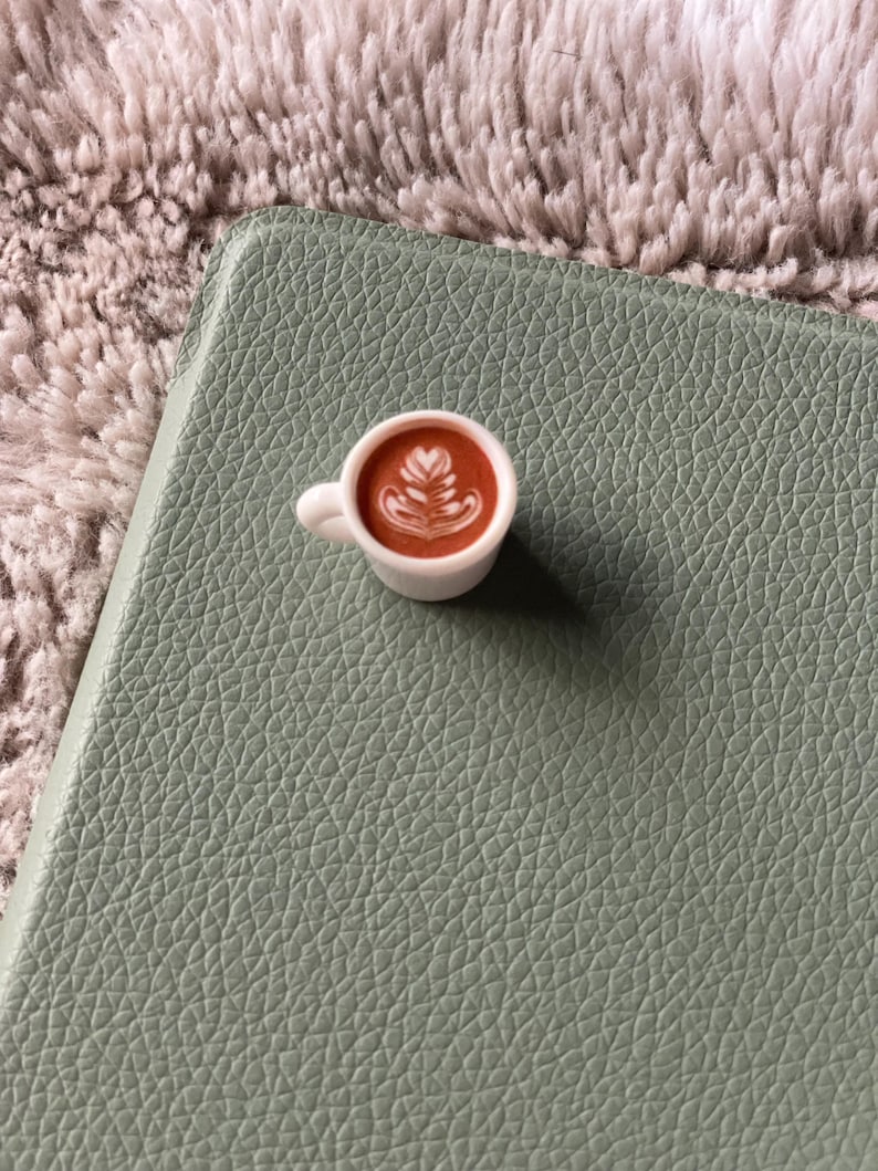Pode incluir: Uma pequena x&iacute;cara de caf&eacute; branca com um design de arte latte, colocada sobre uma superf&iacute;cie texturizada verde s&aacute;lvia. O caf&eacute; &eacute; marrom e a arte latte &eacute; branca. O fundo &eacute; um material macio e difuso, marrom claro.