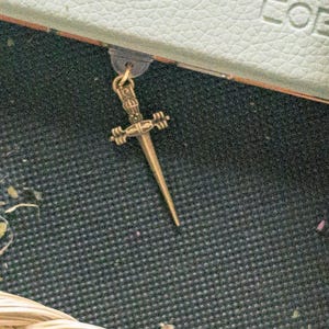 May include: A gold-colored sword bookmark with intricate details. The bookmark is attached to a book with a brown leather cover.